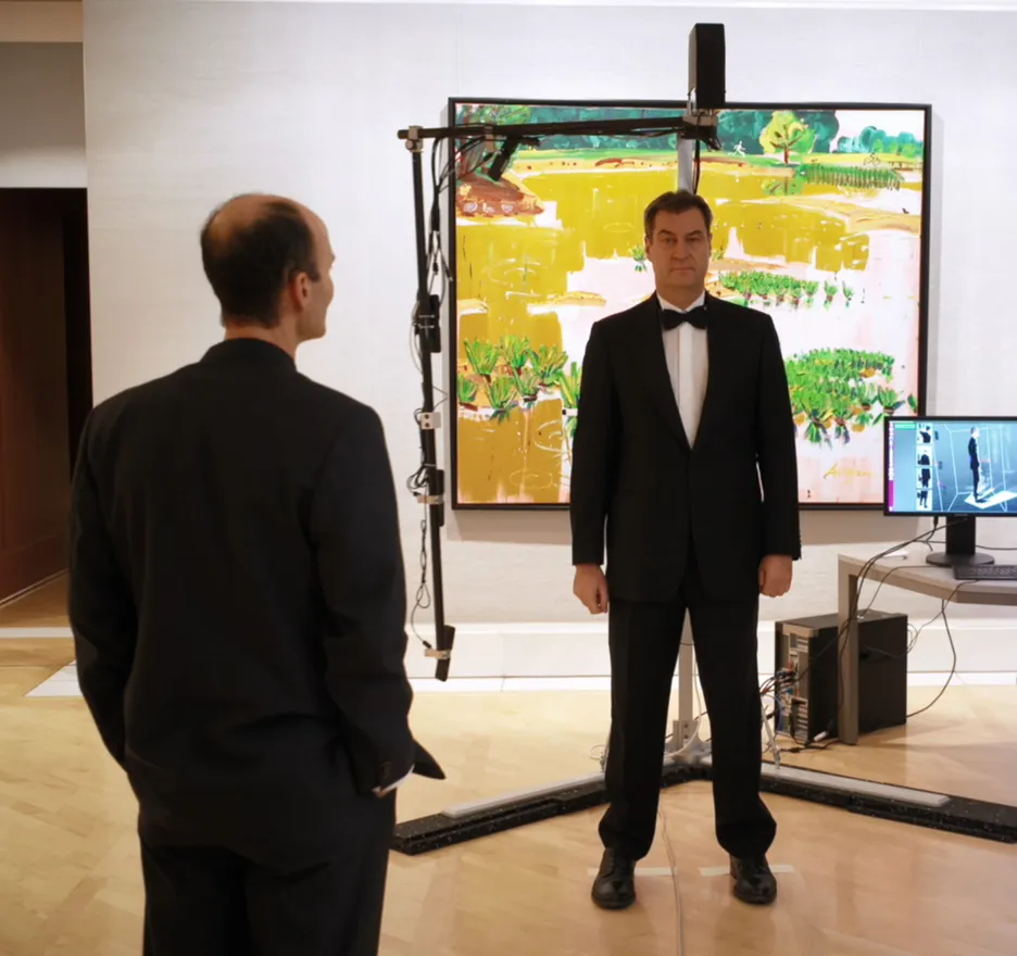Markus Söder is standing in the experimental setup, while Prof. Daniel Cremers is looking at him. A kind of scanner is located above Markus Söder's head, held by a linkage. In the background you can see a computer monitor that shows a human silhouette.